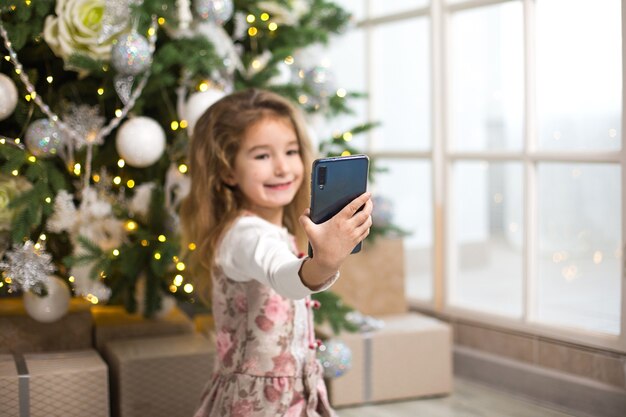 Little girl with Christmas tree takes photos of herself with smartphone