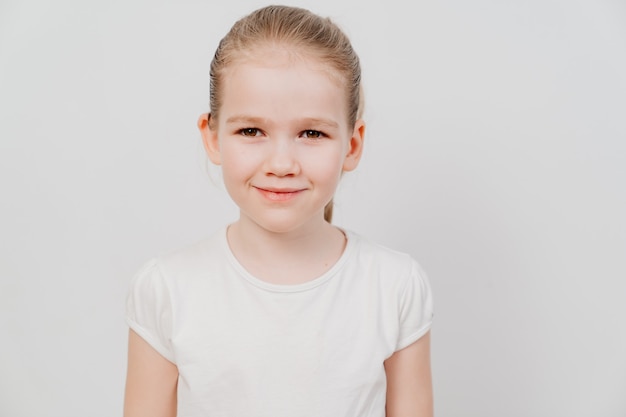 A little girl with collected hair in a white T-shirt stands on a white surface and smiles.