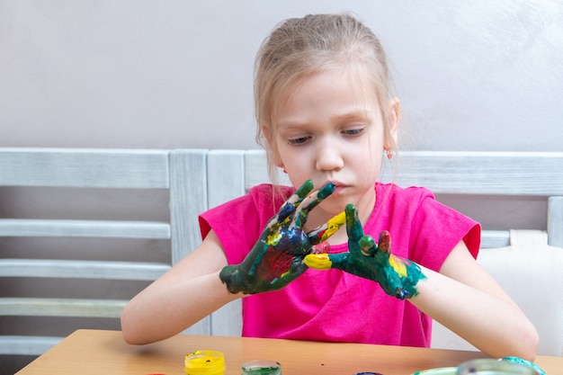 Little girl with colorful painted hands. A concentrated girl smears paints on her hands and looks at the result.