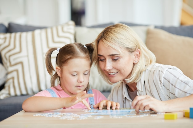 Little girl with down syndrome collecting puzzles at the table with the help of her mother they are in the room at home