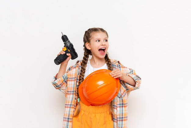 A little girl with an electric screwdriver in a protective orange construction helmet smiles broadly on a white isolated background The concept of renovation in the room of a teenage girl