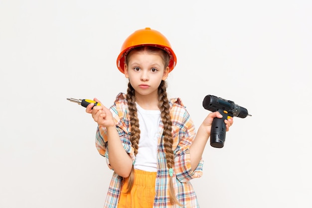 A little girl with an electric screwdriver in a protective orange construction helmet smiles broadly on a white isolated background The concept of renovation in the room of a teenage girl