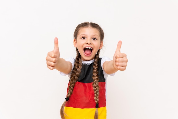 A little girl with a German flag on a Tshirt with long hair in pigtails gives a thumbs up and smiles broadly on a white isolated background Language courses