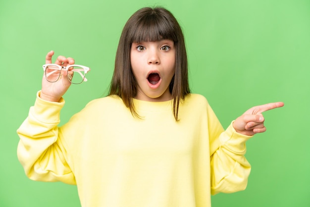 Little girl with glasses over isolated chroma key background surprised and pointing side