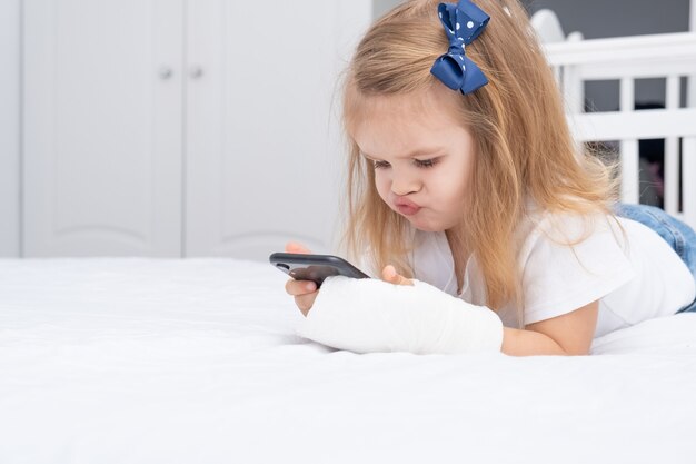 Little girl with hand in cast laying in bed using smartphone, watching cartoon or education video.