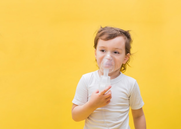 A little girl with an inhaler on a yellow space with copy space. Cute blonde girl with a mask on her face. Looking off to the side.