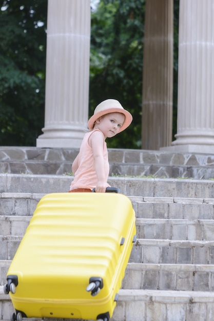 A little girl with light waves understands the steps of a large yellow suitcase. A beautiful child in a straw hat.