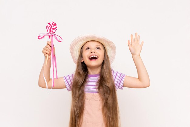 A little girl with a lollipop shows up on a white isolated background A child in a summer jumpsuit and a straw hat eats caramel