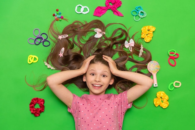 A little girl with long hair lies among elastic bands and hair clips A teenage girl with a comb Accessories for children's hairstyles Green isolated background