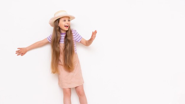 A little girl with long hair in a summer sundress and a straw hat leans against a white walls and looks at your advertisement smilingCopy space