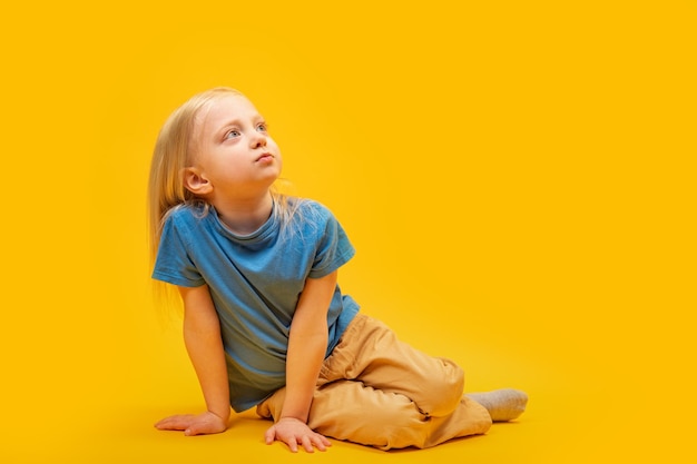 Little girl with loose white hair in blue Tshirt and yellow pants sits on the floor in the studio on yellow background