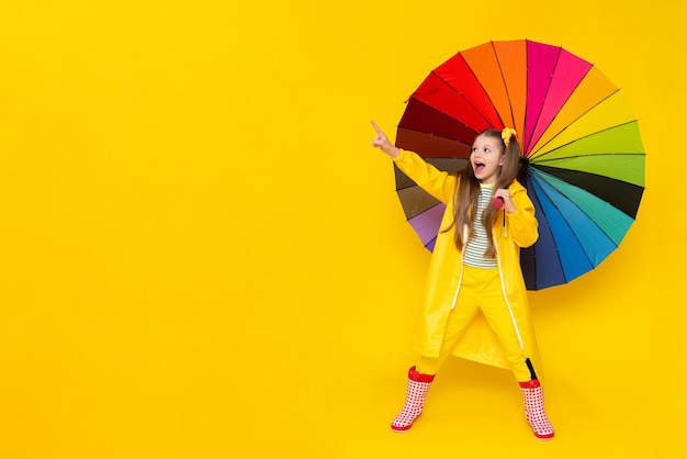 A little girl with a multicolored umbrella in a raincoat and rubber boots on a yellow isolated background Rainy spring weather and puddles Copy space