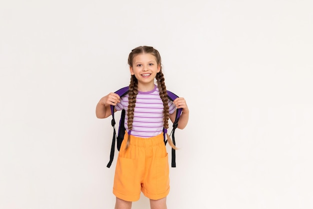 A little girl with pigtails and shorts with a school backpack on her back on a white isolated background Preparatory summer courses for children