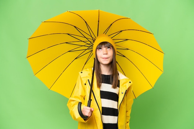 Little girl with rainproof coat and umbrella over isolated chroma key background and looking up