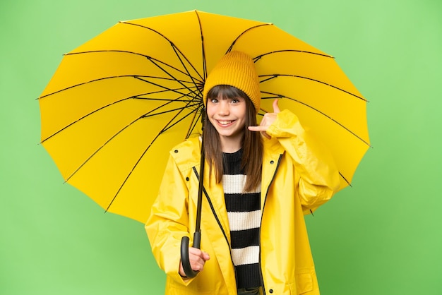 Little girl with rainproof coat and umbrella over isolated chroma key background making phone gesture Call me back sign