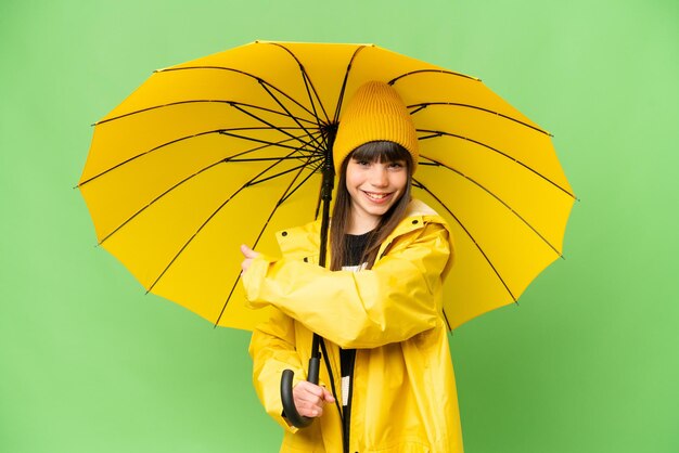 Little girl with rainproof coat and umbrella over isolated chroma key background pointing back