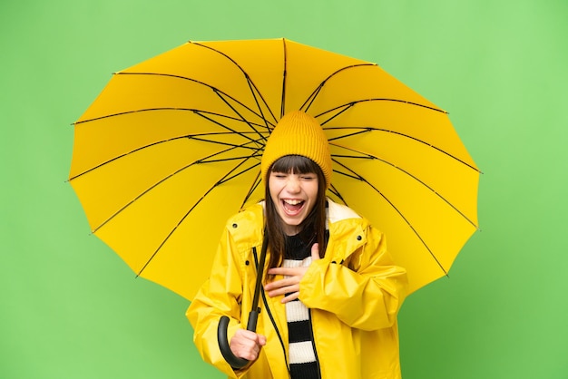 Little girl with rainproof coat and umbrella over isolated chroma key background smiling a lot