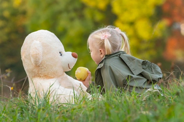 Little girl with two tails plays with teddy bear on grass near autumn forest Fairhaired child in nature with soft toy