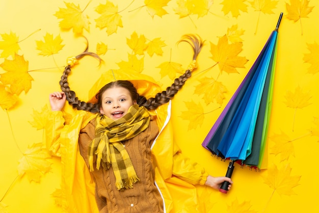 A little girl with an umbrella is lying on autumn yellow maple leaves Funny baby with pigtails in a brown sweater and raincoats