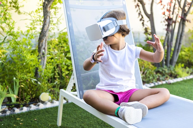 Little girl with virtual reality glasses looking to the left sitting on a sun lounger in the garden of her house Metaverse VR game digital and simulation concept