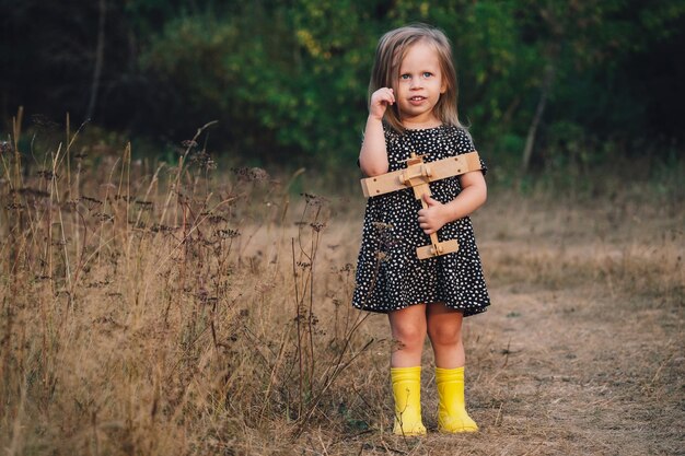 A little girl with a wooden airplane stands in a clearing and looks thoughtfully Sincerely not childish emotions Happy childhood Outdoor games