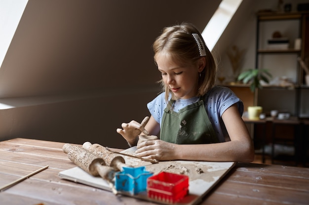 Little girl works with clay at the table, kid in workshop. Lesson at the art school. Young master of folk crafts, pleasant hobby, happy childhood