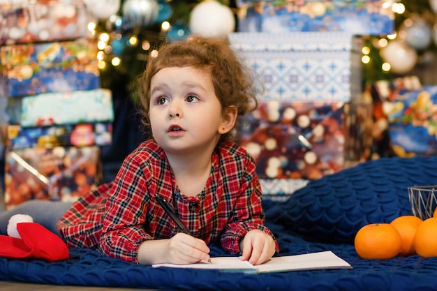 Little girl writing letter to santa. child dream near christmas tree about wish, gift on new year