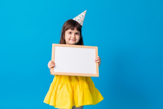 Little girl in a yellow dress on a blue background holds a clean white board with a place for text