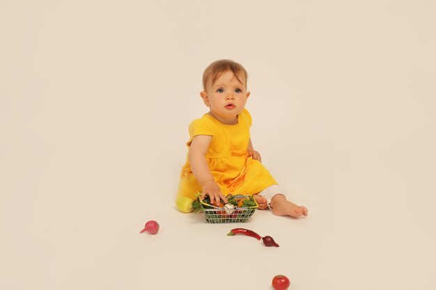 little girl in  yellow dress is sitting on a white background next to a small basket with vegetables