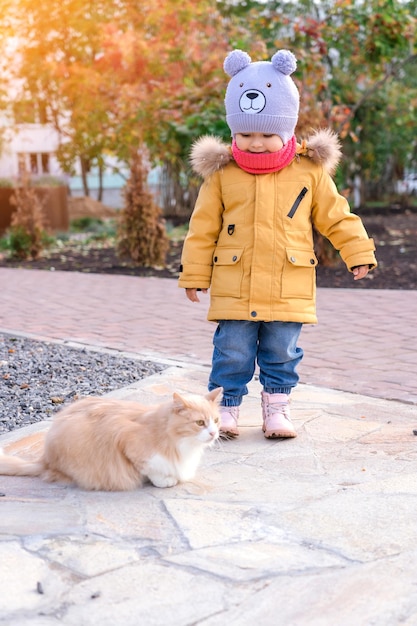 A little girl in a yellow jacket plays with a red cat on a walk in autumn