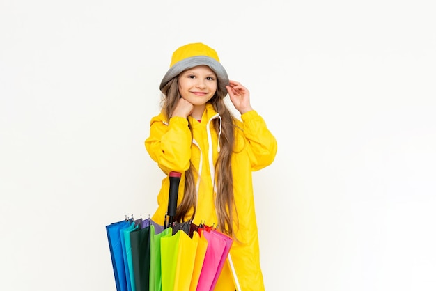 A little girl in a yellow raincoat and panama hat on a white isolated background leans on a multicolored rainbow umbrella