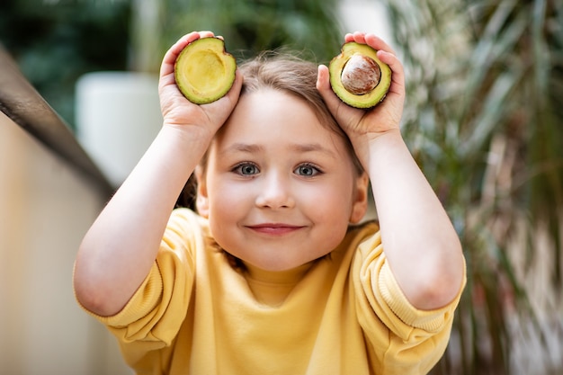 Little girl in yellow sweatshirt holding cut avocado, healthy eating concept.