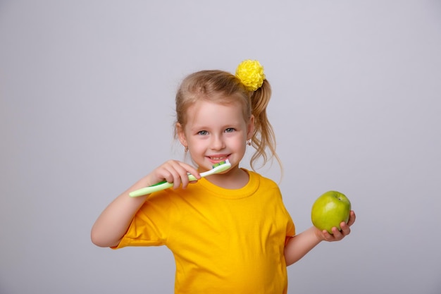 A little girl in a yellow Tshirt holds a toothbrush and an apple on a white background