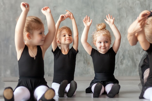 Little girls ballerina in black dresses belk tights and pointe shoes sit on the floor and hold hands over their head for warmup before teaching ballet in a dancing dark studio