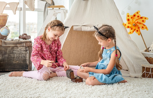 Little girls playing with small treasure box filled with jewels sitting on floor in light room on wigwam