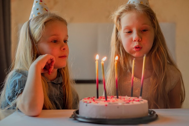 Little girls sisters twins happily blew out the candles on the birthday cake at home festive cap child birthday during illness quarantine isolation