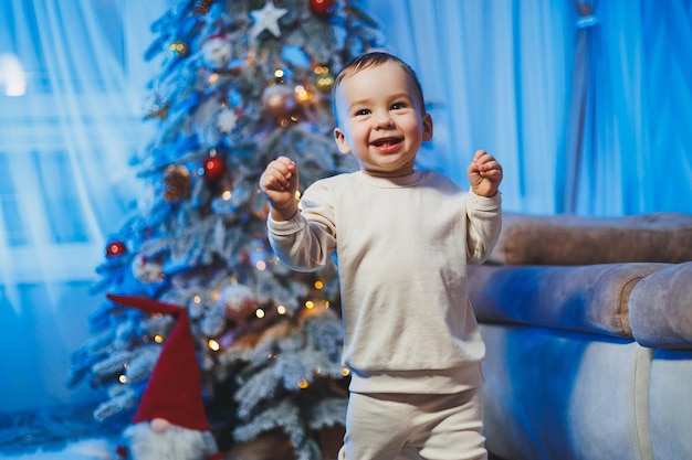 Little handsome oneyearold boy dressed in a beige suit on the background of a Christmas tree in a home environment Children's emotions before celebrating the New Year holiday