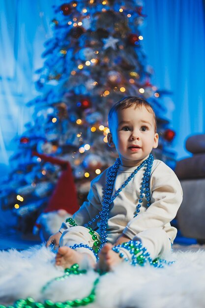 Little handsome oneyearold boy dressed in a beige suit on the background of a Christmas tree in a home environment Children's emotions before celebrating the New Year holiday