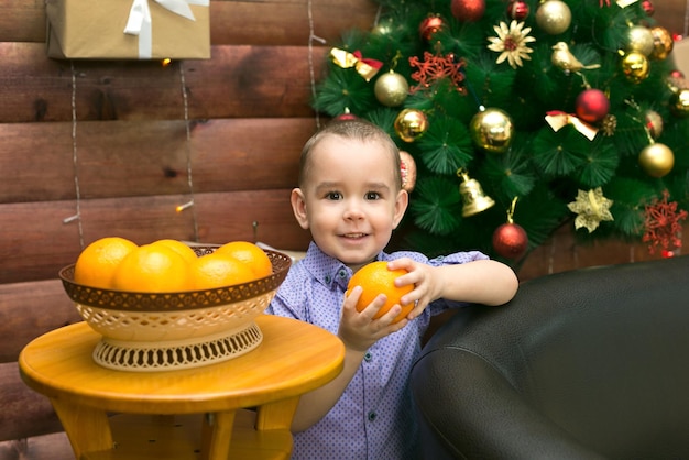 A little happy boy at the Christmas tree holds bright juicy oranges in anticipation of the new year