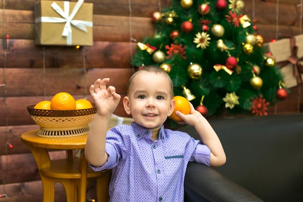 A little happy boy at the Christmas tree holds bright juicy oranges in anticipation of the new year