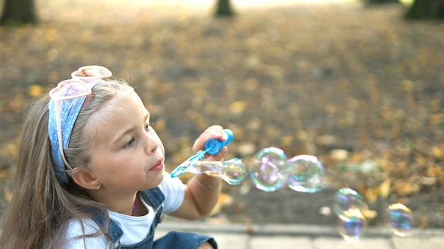 Little happy child girl blowing soap bubbles outside in green park. Outdoor summer activities concept.