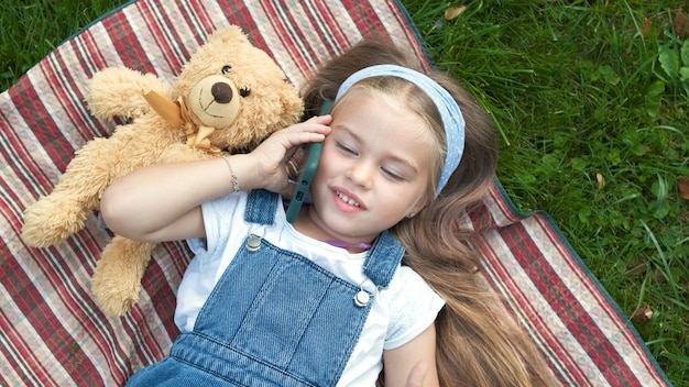 Little happy child girl laying on a blanket on green lawn in summer with her teddy bear talking on mobile phone