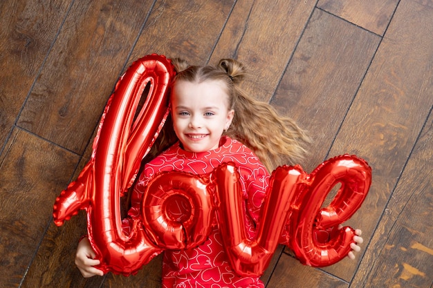 A little happy child girl in red pajamas with a big balloon with the inscription Love lies on a dark brown wooden background on the floor and laughs the concept of Valentines day a place for text