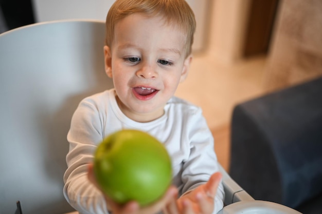 Little happy cute baby toddler boy blonde sitting on baby chair playing with apple baby facial