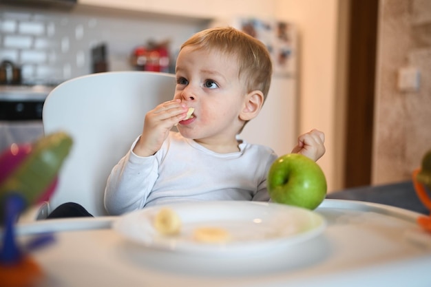 Little happy cute baby toddler boy blonde sitting on baby chair playing with apple baby facial