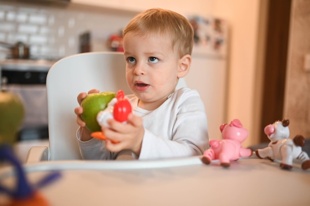 Little happy cute baby toddler boy blonde sitting on baby chair playing with apple baby facial