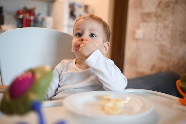 Little happy cute baby toddler boy blonde sitting on baby chair playing with banana baby facial