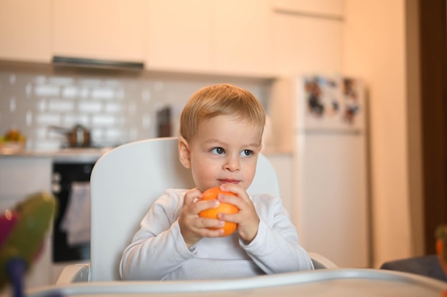 Little happy cute baby toddler boy blonde sitting on baby chair playing with orange baby facial