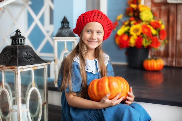 Little happy girl holding pumpkin and smiling sitting on house porch and waiting to trick or treat