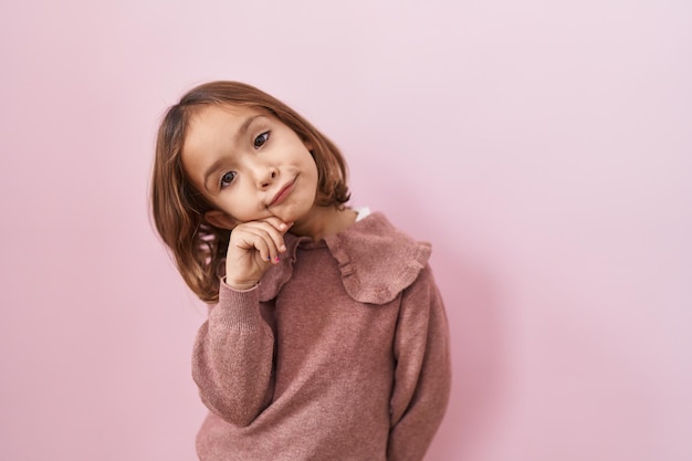 Little hispanic girl standing over pink background serious face thinking about question with hand on chin thoughtful about confusing idea